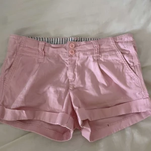 Ljusrosa bomullsshorts med uppvik - Snygga ljusrosa shorts i bomull med uppvikta benslut och två knappar framtill. De har bälteshällor, sidofickor och dekorativa bakfickor. Perfekta för varma dagar och en chill look.