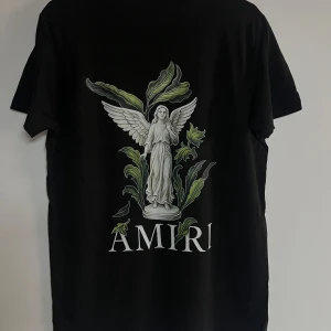 Svart AMIRI t-shirt med ängeltryck - Svart t-shirt från AMIRI med stor grafisk ängel och gröna blad på ryggen samt AMIRI-logga. Framsidan har en mindre logga med färgstänk. Klassisk passform och rund hals. Perfekt för dig som gillar streetwear med konstnärlig vibe.