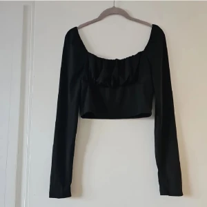 Svart croppad topp från SHEIN - Svart croppad topp från SHEIN med långa ärmar och snygg rynkad detalj framtill. Toppen har en bred, fyrkantig urringning och är tillverkad i ett mjukt, lätt material som känns skönt mot huden. Perfekt för dig som gillar trendiga och stilrena plagg.