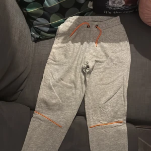 Grå mjukisbyxor med orange snören - Snygga grå mjukisbyxor med orange dragsko i midjan och orange detaljer vid benen. Byxorna har en relaxed fit och är perfekta för chill dagar. Tillverkade i mjukt bomullsmaterial för extra komfort.