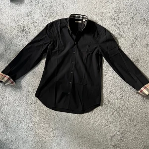 Burberry skjorta (Svart) - Svart skjorta från Burberry med klassiska rutiga detaljer på insidan av kragen och på ärmarna. Den ser helt ny ut och man ser inga täcken på användning. Självklart är den 100% äkta