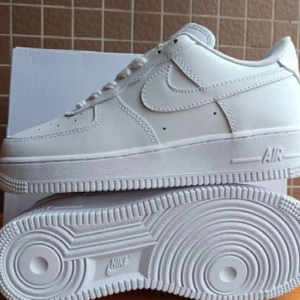 Nike Air Force 1 vita sneakers - Klassiska Nike Air Force 1 sneakers i helvitt skinn med perforerad tå, rund tå och platt sula. Snygg och tidlös modell med snörning och diskret Nike-logga på sidan. Perfekta för dig som vill ha en clean och stilren look.