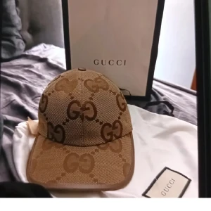 Gucci beige keps med GG-logga - Snygg beige keps från Gucci med klassiskt GG-monogram i brunt över hela kepsen. Kepsen har böjd skärm och justerbar baksida. Perfekt accessoar för att lyfta din outfit med lyxig känsla.