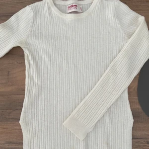 Vit ribbad stickad tröja från Dobber  - Vit ribbstickad tröja från Dobber i storlek XS. Tröjan har rund halsringning, långa ärmar och en snygg metallbricka med logga nertill. Den har även slits som man kan se på bilderna.