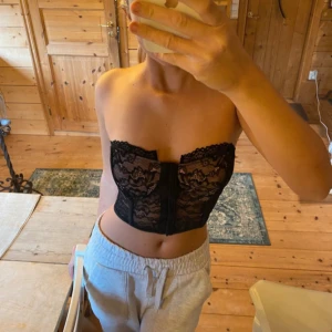 Svart spetskorsett med knäppning  - Säljer en svart korsett-topp i spets med snygg knäppning framtill. Toppen är axelbandslös och har fina spetsdetaljer både fram och bak. Perfekt för dig som vill ha en trendig och feminin look.
