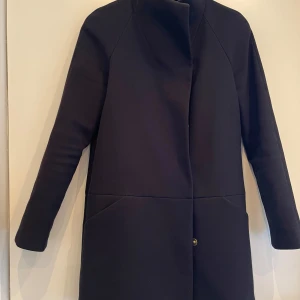 Svart kappa från Mango Suit S - Stilren svart kappa från Mango Suit i storlek S. Jackan har dold dragkedja och tryckknappar i guld, samt två sneda fickor framtill. Klassisk rak modell med lång ärm och hög krage. Perfekt för dig som gillar minimalistisk och clean stil. En knapp saknas samt en öppen söm på insidan av ens ärmen. 