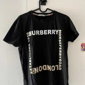 Svart Burberry t-shirt med tryck - Svart t-shirt från Burberry med stort vitt och beige tryck framtill och broderad logga på ärmen. Klassisk rund halsringning och korta ärmar. Snygg och stilren design med London Horseferry Road-tryck. Perfekt för dig som gillar exklusiva streetwear-vibbar.