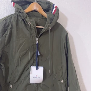 Olivgrön vindjacka från Moncler - Snygg olivgrön vindjacka från Moncler med huva och dragkedja framtill. Jackan har snörning i huvan och Moncler-logga på insidan. Detaljer med röd, vit och blå rand på huvan ger en sportig vibe. Perfekt för dig som gillar stilren och funktionell design.
