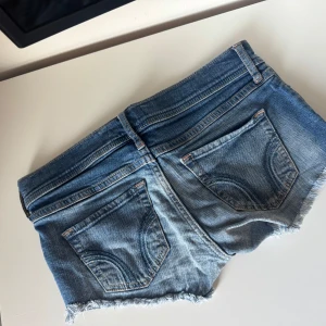 Blå jeansshorts från Hollister - Snygga blå jeansshorts från Hollister med fransig kant och klassiska bakfickor. Modellen har låg midja och slitna detaljer för en avslappnad vibe. Perfekta för varma dagar och festivalhäng. Ikonisk Hollister-logga på knappen och etiketten.
