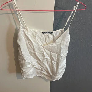 Vit linnetopp från Brandy Melville - Säljer en stilren vit linnetopp från Brandy Melville med tunna justerbara axelband och lätt glansig yta. Toppen har en enkel och clean look, perfekt att matcha med jeans eller kjol. Materialet känns mjukt och svalt mot huden.