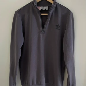 Grå half zip tröja från Adidas - Snygg grå långärmad tröja från Adidas med svart logga på bröstet och half zip-dragkedja. Tröjan har en enkel och clean design med hög krage och är tillverkad i mjuk bomullsblandning. Perfekt för chill dagar eller när du vill ha en sportig vibe.