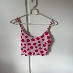 Jordgubbsmönstrad rosa linnetopp XS - Supersöt croppad rosa linnetopp från SHEIN med jordgubbsmönster över hela toppen. Tunt och mjukt material, smala axelband och vågig kant nertill. Perfekt för dig som gillar färgglada och lekfulla plagg!
