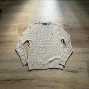 Beige kabelstickad tröja Polo Ralph Lauren - Kabelstickad beige tröja från Polo Ralph Lauren med klassisk rund hals och broderad logga på bröstet. Tröjan är i bomull och har ribbade muddar vid ärmslut och nederkant. Perfekt för lager-på-lager under hösten och vintern. Ny skick, M men funkar även som L