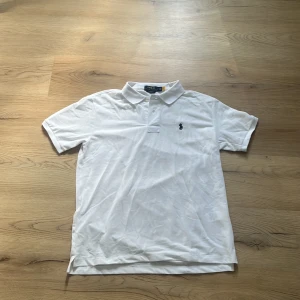 Vit pikétröja från Polo Ralph Lauren - Klassisk vit pikétröja från Polo Ralph Lauren i modellen Custom Slim Fit. Tröjan har korta ärmar, krage och knäppning framtill samt den ikoniska marin blåa broderade loggan på bröstet. Tillverkad i bomull för en skön och luftig känsla. Ny skick, sitter som M men funkar även som L
