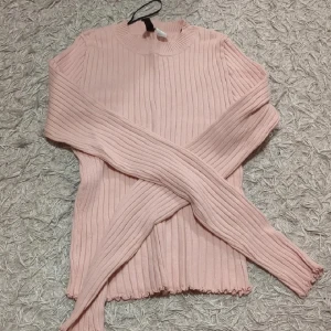 Ljusrosa ribbad stickad tröja - Supersöt ljusrosa stickad tröja med ribbat mönster och långa ärmar. Tröjan har en högre rund halsringning och vågig kant nertill för en extra detalj. Perfekt att styla med jeans eller kjol för en chill look.