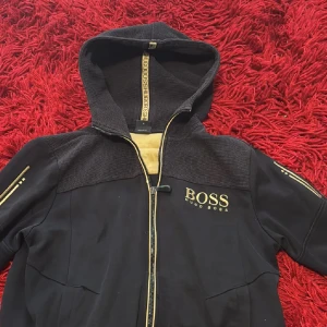 Svart hoodiejacka från Hugo Boss - Svart hoodiejacka från Hugo Boss med guldiga detaljer och dragkedja. Jackan har huva, guldiga logotyper och detaljer på ärmarna samt ribbade partier upptill. Perfekt för dig som vill ha en stilren och sportig look med exklusiv känsla.