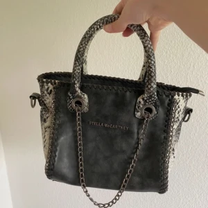 Stella McCartney grå handväska med kedja - Snygg svart handväska från Stella McCartney med coola ormmönstrade handtag och detaljer. Väskan har silverfärgad kedja. Fint skick