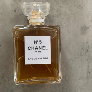 Chanel No 5 Eau de Parfum 100ml - Klassisk Chanel No 5 Eau de Parfum i 100 ml flaska. Flaskan är rektangulär i genomskinligt glas med en elegant, transparent kristalliknande kork. Parfymen har en gyllene bärnstensfärg och ikonisk vit etikett med svart text. Perfekt för dig som gillar tidlös och lyxig doft.  ‼️‼️OBS‼️‼️Det finns ingen kartong 