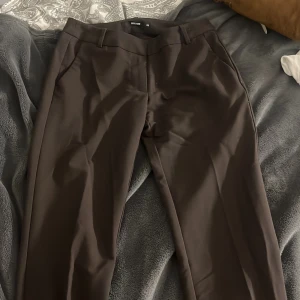 Bruna kostymbyxor lågmidjade  bootcut - Snygga bruna kostymbyxor med klassisk bootcut passform och pressveck. Byxorna har diskreta sidfickor och hällor för bälte. Perfekta för en clean och stilren look. Materialet känns mjukt och följsamt, troligtvis polyesterblandning. Mina har ingen knapp på för den har gått av men det finns en extra knapp på sidan av byxorna så man kan lägga till kanppen men den är inte nödvändig, använt dem ksk 4 gånger dem är i nyskick 