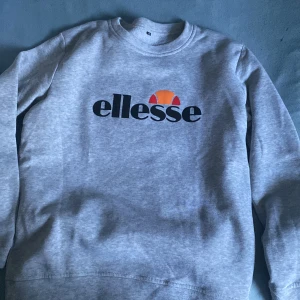 Grå sweatshirt från Ellesse stl M - Säljer en grå sweatshirt från Ellesse med klassisk logga i svart, orange och rött framtill. Tröjan har rund halsringning och långa ärmar. Perfekt för en chill och sportig look.