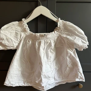 Vit blus med puffärm från Pimkie - Supersöt vit blus från Pimkie med broderade blommor och puffiga korta ärmar. Smockad resår vid halsringning och ärmslut ger en romantisk vibe. Perfekt för dig som gillar en luftig och feminin stil. Tillverkad i bomull för en skön känsla.