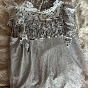 Vit blus med volang från Holly & Whyte - Supersöt vit blus från Holly & Whyte med broderade detaljer och volang längs axlarna. Blusen är ärmlös och har en romantisk känsla med spets och små hålbroderier upptill. Perfekt för dig som gillar en feminin och luftig stil.