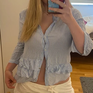 Blåvit randig volangskjorta - Säljer en blåvit randig skjorta med volangdetaljer nedtill och knappar. Saknar en knapp på ena ärmen längst ner därav priset. 