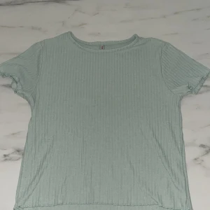 Ljusgrön-turkos ribbad t-shirt från ONLY - Snygg ljusgrön t-shirt från ONLY med ribbad struktur och korta ärmar. T-shirten har en enkel rund halsringning och är tillverkad i mjukt material som känns skönt mot huden. Perfekt för en chill och avslappnad stil.