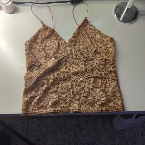 Beige spetslinne från NLY Trend - Supersnyggt beige linne från NLY Trend med tunna axelband och v-ringning. Toppens yta är täckt av blommig spets och har en lätt glansig underton. Perfekt för dig som gillar spets detaljer och vill sticka ut lite extra.