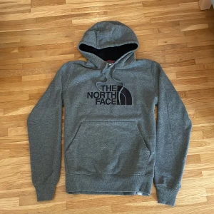 Grå hoodie från The North Face - Säljer en grå hoodie från The North Face med svart logga på bröstet. Tröjan har huva med snörning, stor magficka och långa ärmar. Mjuk insida och klassisk passform, perfekt för chill dagar eller när du vill vara extra bekväm.