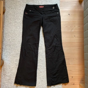 Svarta bootcut byxor från Gant - Svarta byxor från Gant i bootcut-modell med låg midja. Snygga detaljer med silverfärgade knappar och fickor både fram och bak. Lite tunnare i materialet. 