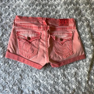 Rosa jeansshorts från True Religion - Snygga rosa jeansshorts från True Religion med klassiska bakfickor och kontrastsömmar. Modellen har uppvikta ben, fem fickor och metallknappar. Perfekt för dig som vill ha en cool och avslappnad look i sommar. Lågmidjade och något slitna vid bakfickorna. 