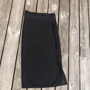 Svart långkjol från 157 - Svart långkjol från 157 med stilren design och en liten slits på sidan. Kjolen är gjord i mjukt och stretchigt syntetmaterial som sitter bekvämt och har en clean look. Perfekt för dig som gillar minimalistiska plagg med enkel passform.