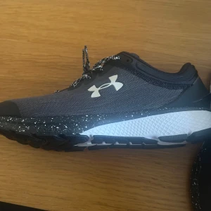 Under Armour Charged Escape sneakers - Snygga svarta och grå Under Armour Charged Escape sneakers med vit logga på sidan och hälen. Skorna har en platt sula med coolt stänkmönster i vitt och svarta/vita snören. Ovandelen är i mesh för bra ventilation och komfort, perfekt för dig som gillar träning och streetstyle.