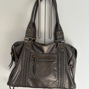 Silvrig handväska med flätade detaljer - Snygg silvrig handväska med flätade detaljer och dekorativa nitar. Väskan har en praktisk framficka med dragkedja och rejäla handtag. Perfekt storlek för att få plats med allt du behöver och ger en cool touch till din outfit.