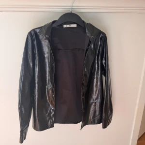 Svart glansig skjorta från NA-KD - Svart skjorta från NA-KD med glansig finish och klassisk krage. Långärmad modell med knappar framtill. Materialet har en skinnliknande look som ger en cool vibe. Perfekt för dig som gillar att sticka ut med din stil.