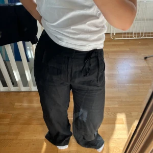 Svarta linnebyxor med snörning - Snygga svarta linnebyxor med avslappnad passform och knytband i midjan. Byxorna har vida ben och är perfekta för dig som gillar en chill och stilren look. Materialet är linne vilket gör dem luftiga och sköna att bära. Passar lika bra till sneakers som till sandaler.
