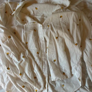 Vit blommig skjorta med knappar - Säljer en vit skjorta med små gula blommor broderade över hela tyget. Skjortan har långa ärmar med resår vid ärmslut och klassisk krage. Stängs med knappar framtill. 