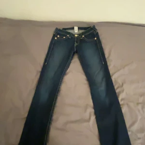 True Religion mörkblå jeans bootcut - Snygga mörkblå jeans från True Religion med klassisk gul kontrastsöm och ikonisk broderad ficka bak. Modellen har bootcut-ben och låg midja, vilket ger en schysst siluett. Jeansen är gjorda i slitstarkt denim och har detaljer som knappar och dragkedja framtill.