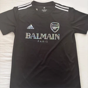 Svart Arsenal x Balmain fotbollströja - Svart fotbollströja från Adidas med Arsenal-logga och Balmain Paris-tryck i holografisk stil. 