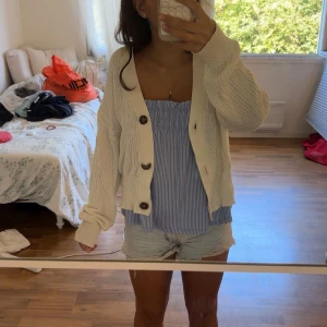 Vit stickad cardigan med knappar - Säljer en vit stickad cardigan jacka med grova bruna knappar och v-ringad hals. Modellen är loose och har långa ärmar med breda muddar. Perfekt att slänga över ett linne. Materialet är mjukt och luftigt stickat. Tyvärr har mellersta knappen gått sönder men det går fortfarande att knäppa 