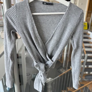 Grå och beige omlottoppar från Zara - Två ribbade omlottoppar från Zara i storlek S, en i ljusgrå och en i beige. Båda har långa ärmar, djup v-ringning och knyt framtill. Perfekt för att skapa en chill och snygg look, och de är gjorda i mjuk bomullsmix som känns skön mot huden.( båda för 110kr ena för 70kr)