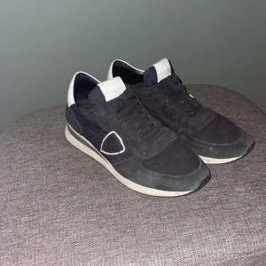 Svarta Philippe Model sneakers - Snygga svarta sneakers från Philippe Model med vita detaljer på häl och plös. Skorna har snörning, rund tå och platt sula. Materialmix av mocka och syntet ger en cool och sportig vibe. Perfekta för dig som gillar stilrena och trendiga sneakers.