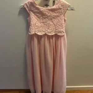 Ljusrosa långklänning med spets och rosett - Superfin ljusrosa långklänning med spets upptill och mjukt, fallande tyg nedtill. Klänningen har ärmlös design och en söt rosett i ryggen som knyts. Spetsen har ett blommigt mönster och kanten är vågig för extra detalj. Perfekt för speciella tillfällen!