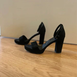 Svarta klackade sandaler med plattform - Snygga svarta sandaler med bred klack och plattform. Skorna har öppen tå och ett brett band över foten samt justerbart spänne vid ankeln. Perfekta för dig som gillar stilrena och höga klackar. Materialet ser ut att vara mocka för en lyxig känsla.