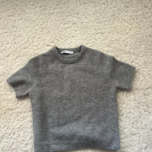 Grå stickad kortärmad tröja från Zara - Säljer en enkel och stilren grå stickad tröja från Zara med korta ärmar och rund hals. Tröjan har ribbade muddar vid ärmslut och nederkant, perfekt för dig som gillar en clean look. Materialet är mjukt och skönt mot huden.