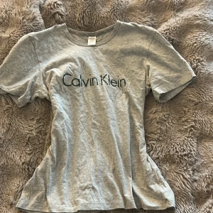 Grå Calvin Klein t-shirt med logga - Snygg grå t-shirt från Calvin Klein med klassisk svart logotyp tryckt på bröstet. Modellen har rund hals och korta ärmar, tillverkad i mjuk bomull som är skön mot huden. Perfekt för chill dagar eller när du vill ha en clean look.