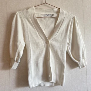 Vit ribbad kofta från NA-KD - Snygg vit kofta från NA-KD i ribbad bomull med v-ringning och två stora knappar framtill. Koftan har puffade trekvartsärmar och en mjuk, lite loose passform. Perfekt att slänga över en topp för en chill och fräsch look.