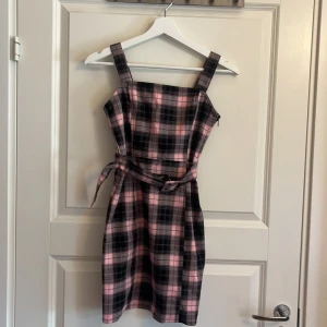 Rutig rosa och svart klänning från H&M💓 - Supersöt kort klänning från H&M i rosa och svart rutigt mönster. Klänningen har breda axelband, bälte i midjan och dragkedja på sidan. Axelbanden är någon centimeter uppsydd men är enkel att få upp. Perfekt för dig som gillar en lekfull och trendig stil. Materialet är mjukt och bekvämt, och den är ärmlös för en fräsch look💓.