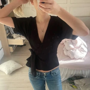 Svart omlottopp med knyt - Snygg svart omlottopp med korta ärmar och v-ringning. Toppen har ett knyt i midjan som ger en cool och feminin siluett. Perfekt att matcha med jeans eller kjol för en trendig look.